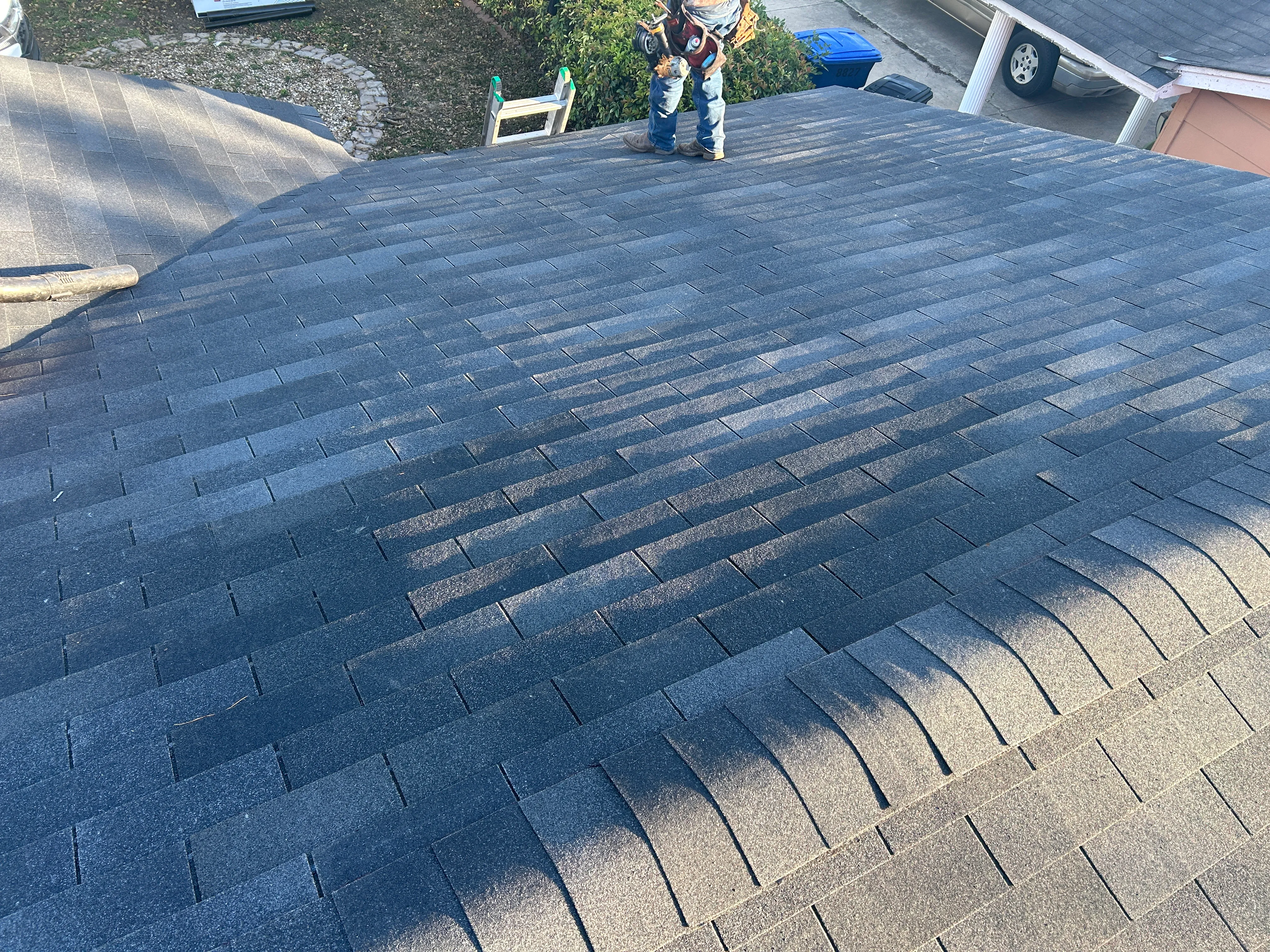 After: completed dark gray asphalt shingle roof showing a valley and ridge lines, with a truck and work trailer visible beyond the property.